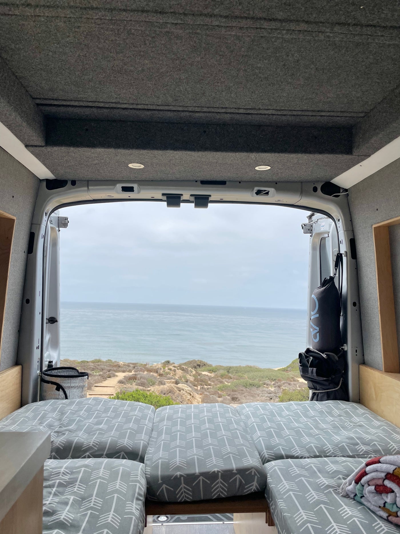 Inside an RV rental, a bed with arrow-patterned cushions faces a panoramic ocean view over a coastal landscape. — photo 3