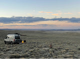 Relaxing RV rental experience: A campervan, glowing campfire, and deer at dusk in an expansive, grassy landscape.