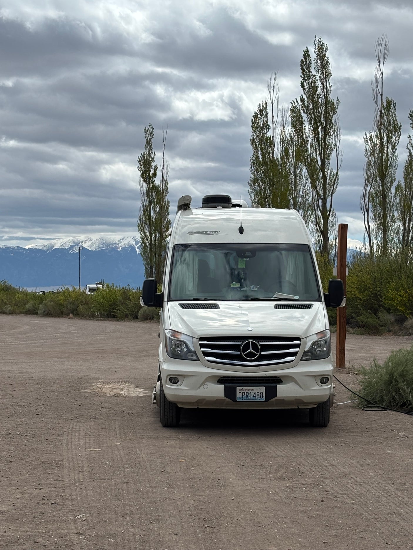 White Mercedes Sprinter RV rental parked on gravel, with snow-capped mountains and tall trees under cloudy skies.