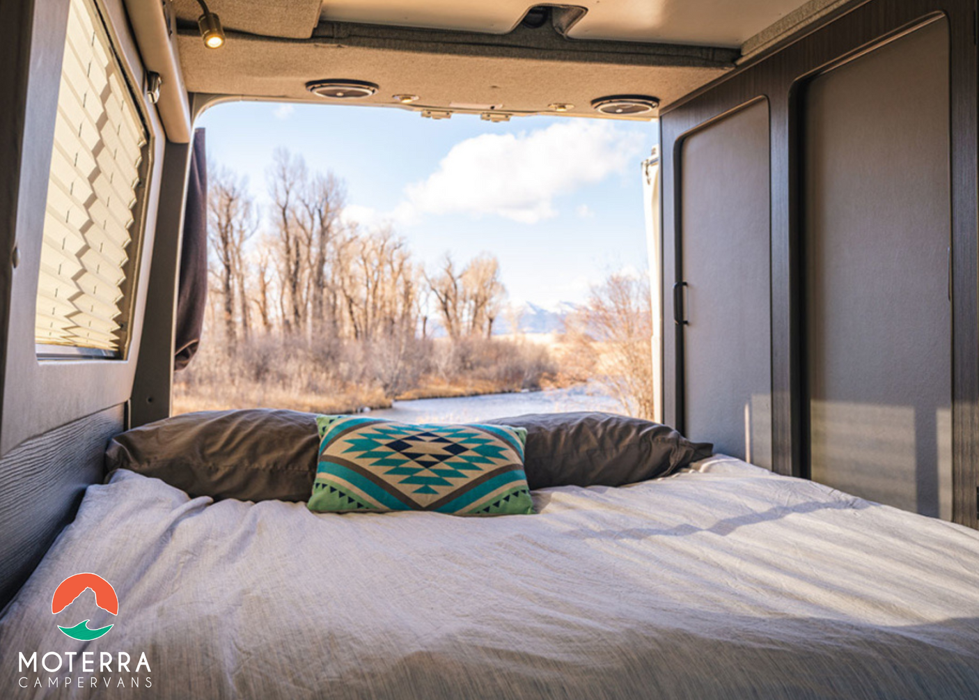 Relax in this cozy RV rental bed with open doors revealing a picturesque river, bare trees, and distant mountains under a blue sky.