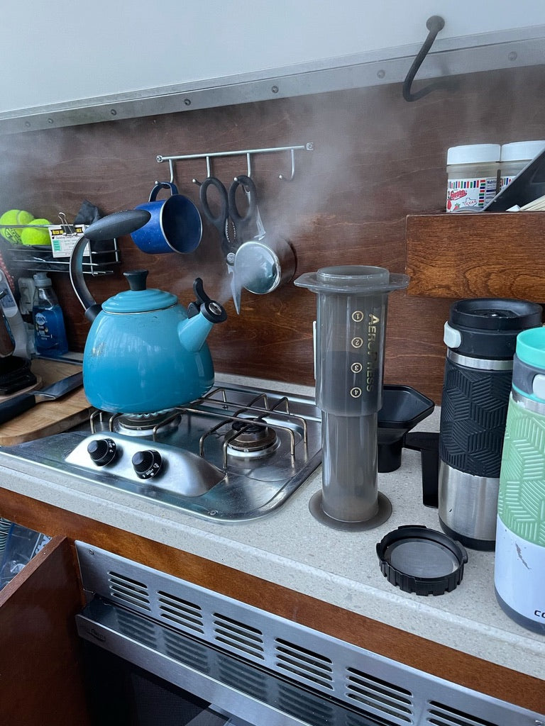 Steaming kettle & AeroPress make coffee in an RV rental kitchen. Compact setup with mugs, ideal for on-the-go travel.