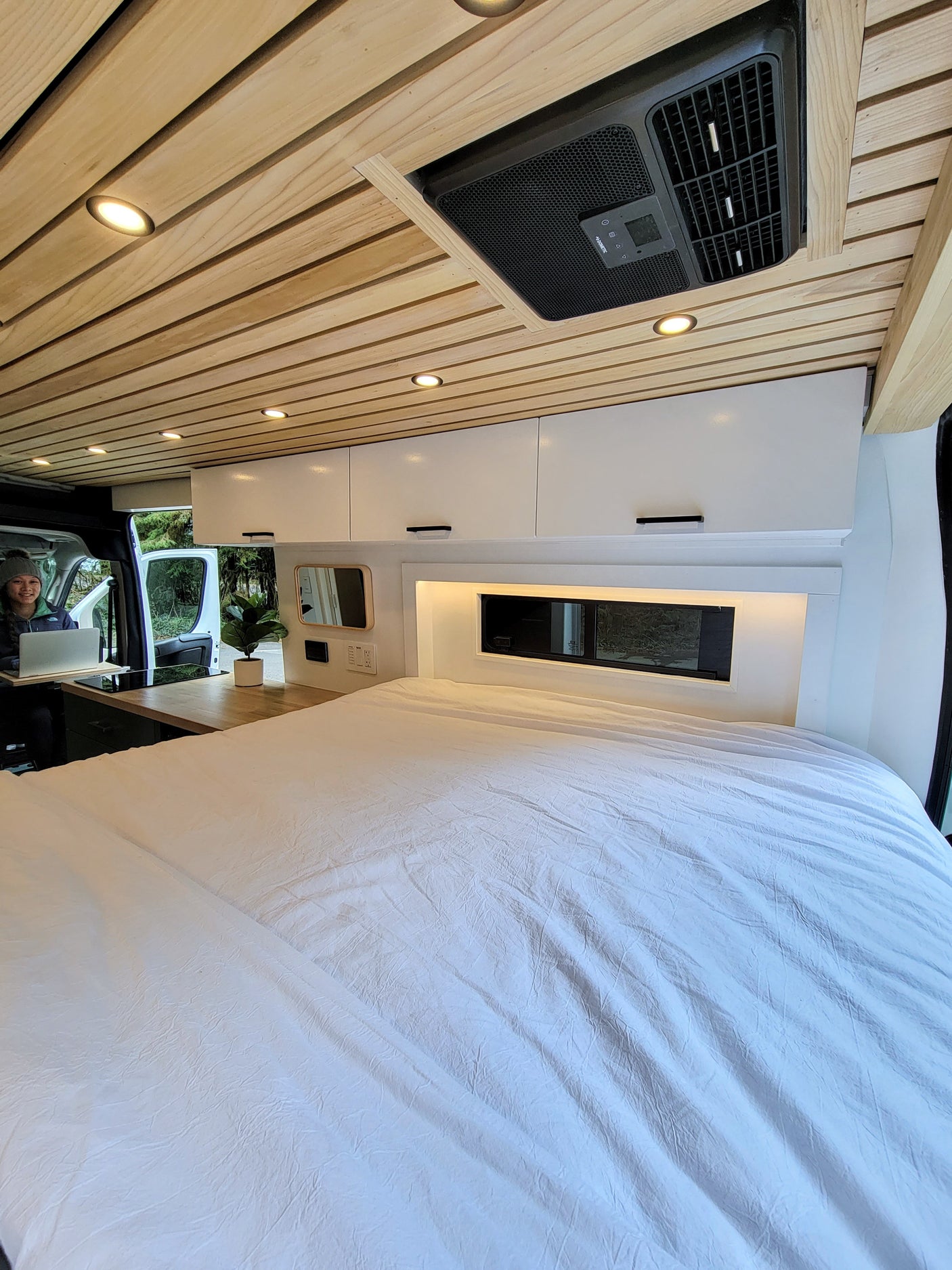 Cozy RV rental interior featuring a wood ceiling, bright lights, and a large bed. A person works at the built-in kitchen/desk area.
