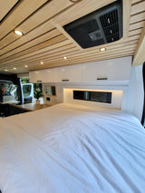 Cozy RV rental interior featuring a wood ceiling, bright lights, and a large bed. A person works at the built-in kitchen/desk area.