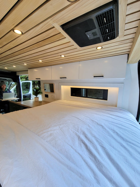Cozy RV rental interior featuring a wood ceiling, bright lights, and a large bed. A person works at the built-in kitchen/desk area.