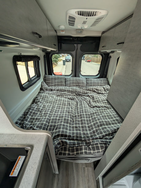 Inside a comfy RV rental: A grey plaid bed, overhead cabinets, and compact kitchen area for mobile travel.