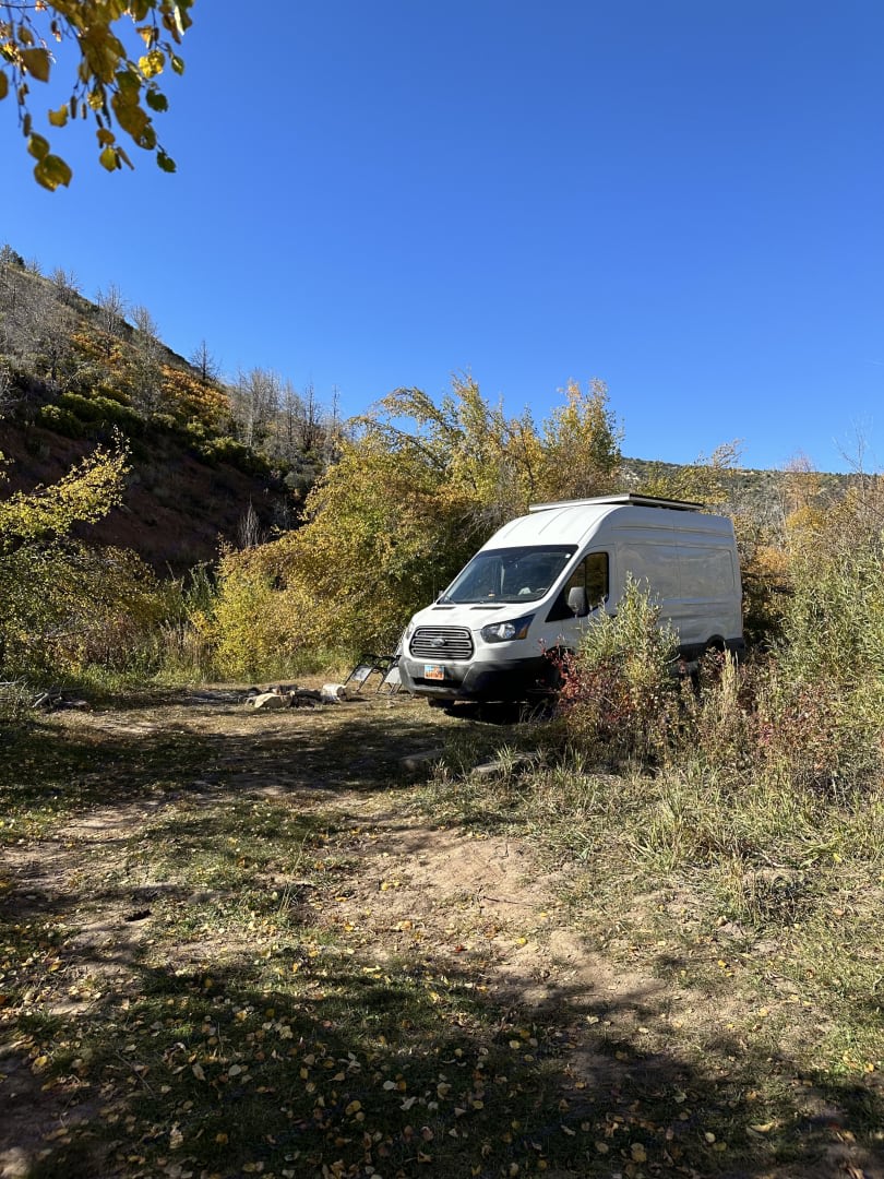 White RV rental camper van at a rustic autumn campsite with a fire pit, surrounded by fall foliage under a clear blue sky.