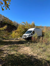 White RV rental camper van at a rustic autumn campsite with a fire pit, surrounded by fall foliage under a clear blue sky.