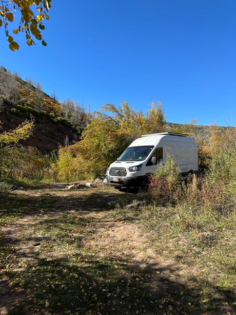 White RV rental camper van at a rustic autumn campsite with a fire pit, surrounded by fall foliage under a clear blue sky.