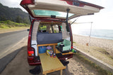 Red RV rental camper van parked at the beach, rear open with bed, pull-out kitchen, table, and awning setup.