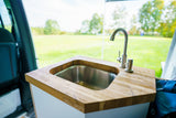 RV rental's compact kitchen with a built-in stainless steel sink, wood counter, and outdoor field view.