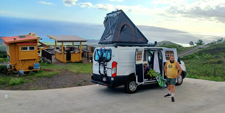 Scenic Maui overlook with a white RV rental van, rooftop tent, and open side door showing fresh fruit. A man stands nearby, ocean view.