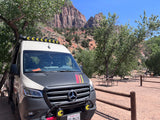 White & black custom RV rental van with stripes, parked in a sunny desert campground with red rock mountains.