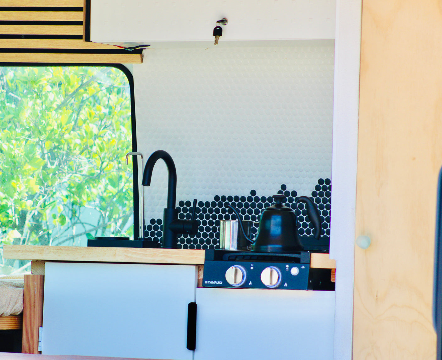 An RV rental's cozy kitchen features a black faucet, penny tile backsplash, stovetop with a kettle, and a leafy view outside.