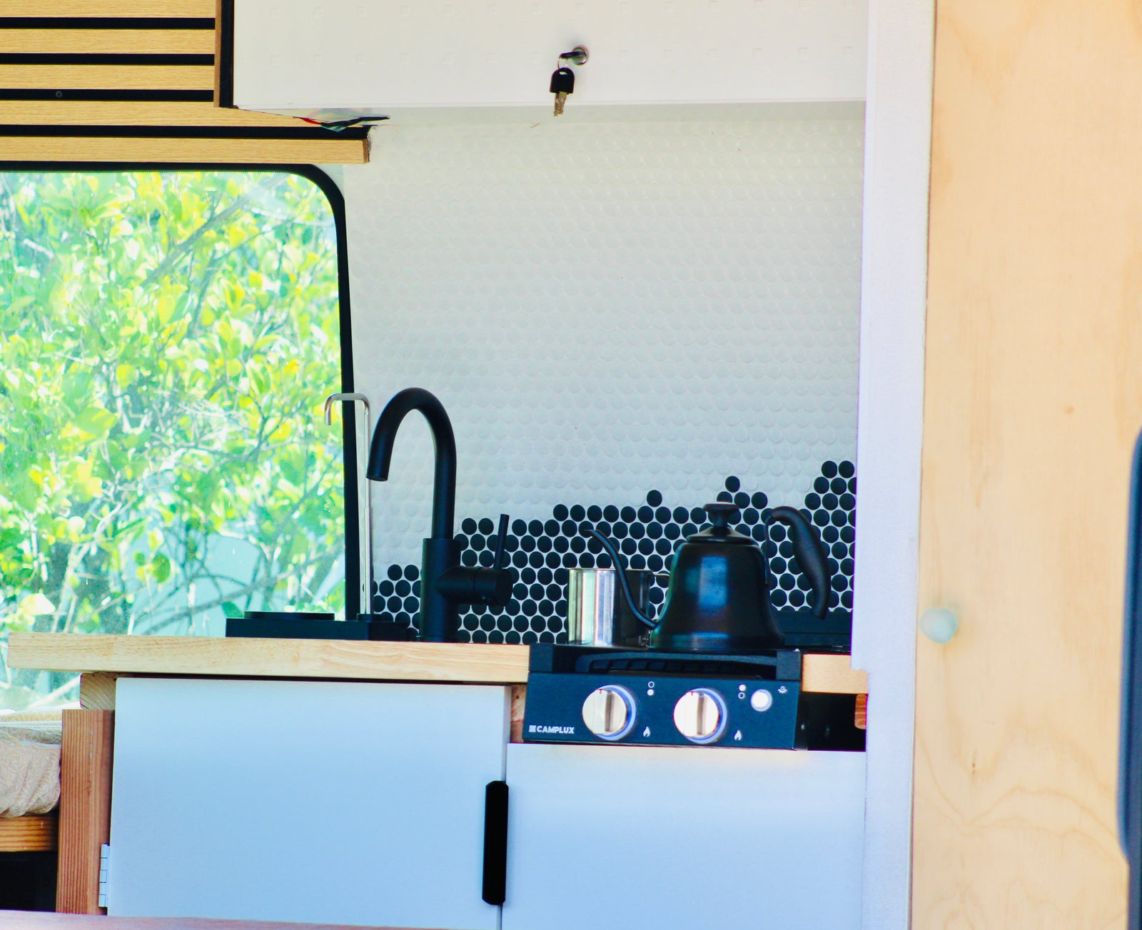 An RV rental's cozy kitchen features a black faucet, penny tile backsplash, stovetop with a kettle, and a leafy view outside.