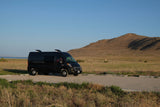 Black RV rental van parked by a lake with a large mountain backdrop, perfect for outdoor adventures.