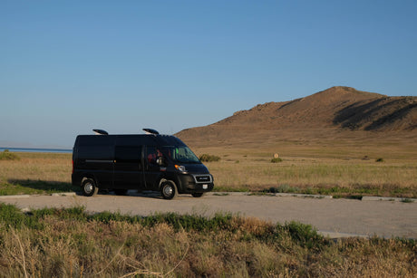Black RV rental van parked by a lake with a large mountain backdrop, perfect for outdoor adventures.