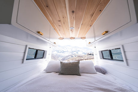 Modern RV rental: a cozy bedroom with overhead cabinets, wooden ceiling, and snowy mountain views.