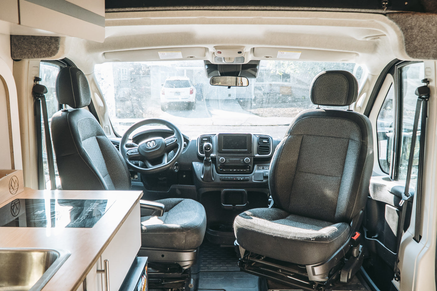 Inside an RV rental camper van: cockpit with driver/passenger seats, dashboard, kitchenette, and a view of a white SUV outside.
