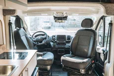 Inside an RV rental camper van: cockpit with driver/passenger seats, dashboard, kitchenette, and a view of a white SUV outside.