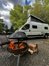 Perfect RV rental camp setup: White camper van, crackling fire pit, and firewood on gravel under a cloudy sky.