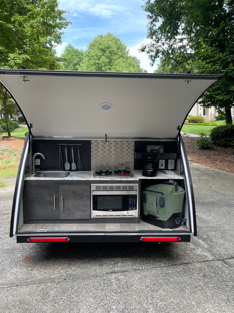 This rv rental features an open-hatch outdoor kitchen with a sink, stovetop, microwave, coffee maker, and cooler setup.