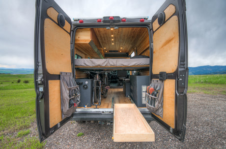 RV rental: Rear view of a custom camper van, doors open, revealing a wood interior, bed, bike storage & pull-out tray in nature.