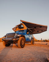 Blue Jeep Wrangler with rooftop tent & awning at sunset in the desert. Ideal rv rental for off-road camping adventures.