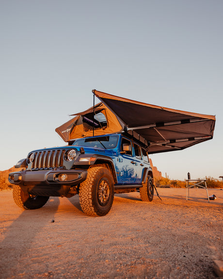 Blue Jeep Wrangler with rooftop tent & awning at sunset in the desert. Ideal rv rental for off-road camping adventures.