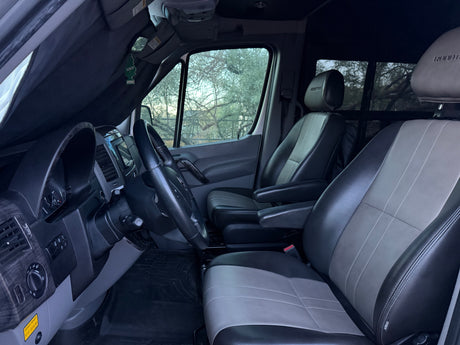 Front cabin of an rv rental showing the dashboard, steering wheel, and custom black & grey leather seats with armrests.