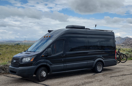 Black Ford Transit RV rental with roof AC, solar panels, and bike rack carrying two bikes, parked on a road with mountains.