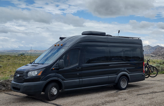 Black Ford Transit RV rental with roof AC, solar panels, and bike rack carrying two bikes, parked on a road with mountains.