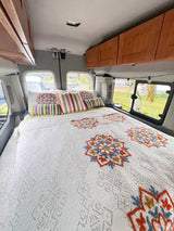 This RV rental features a cozy bedroom with a white quilt, colorful mandala embroidery, striped pillows, and wooden overhead cabinets.