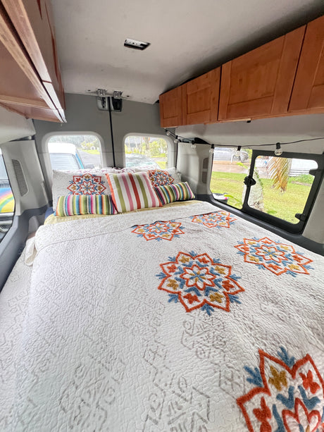 This RV rental features a cozy bedroom with a white quilt, colorful mandala embroidery, striped pillows, and wooden overhead cabinets.