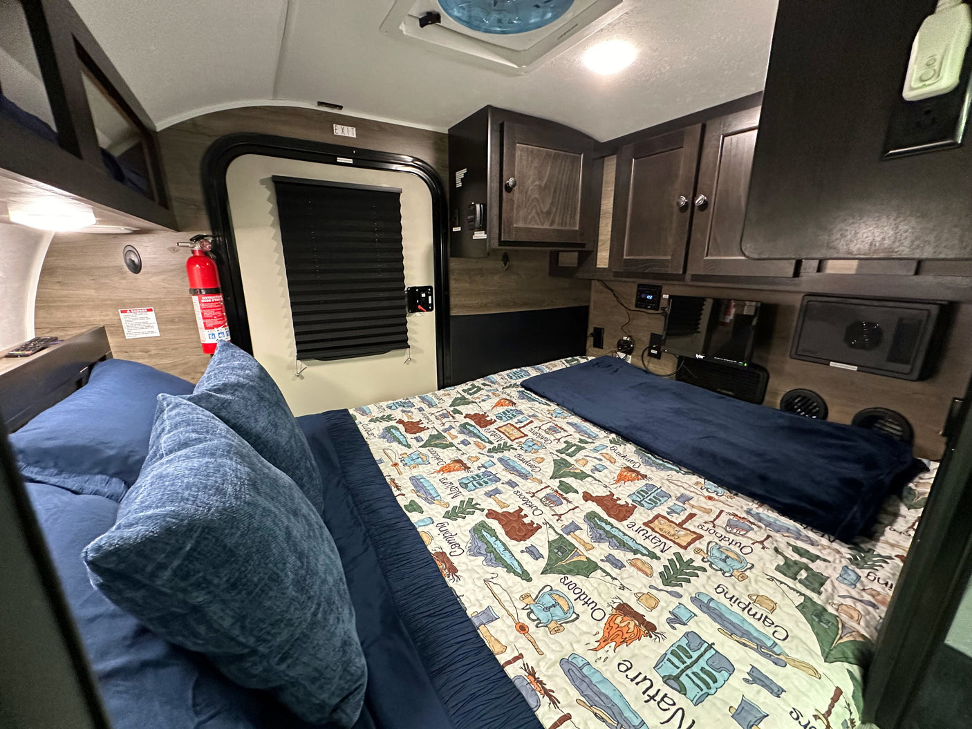 Cozy RV rental sleeping area featuring a nature-themed bed, exit door, fire extinguisher, and overhead storage.