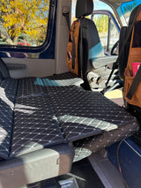 Black quilted mattress fills this RV rental van's interior, showing autumn leaves outside. Ready for adventure!