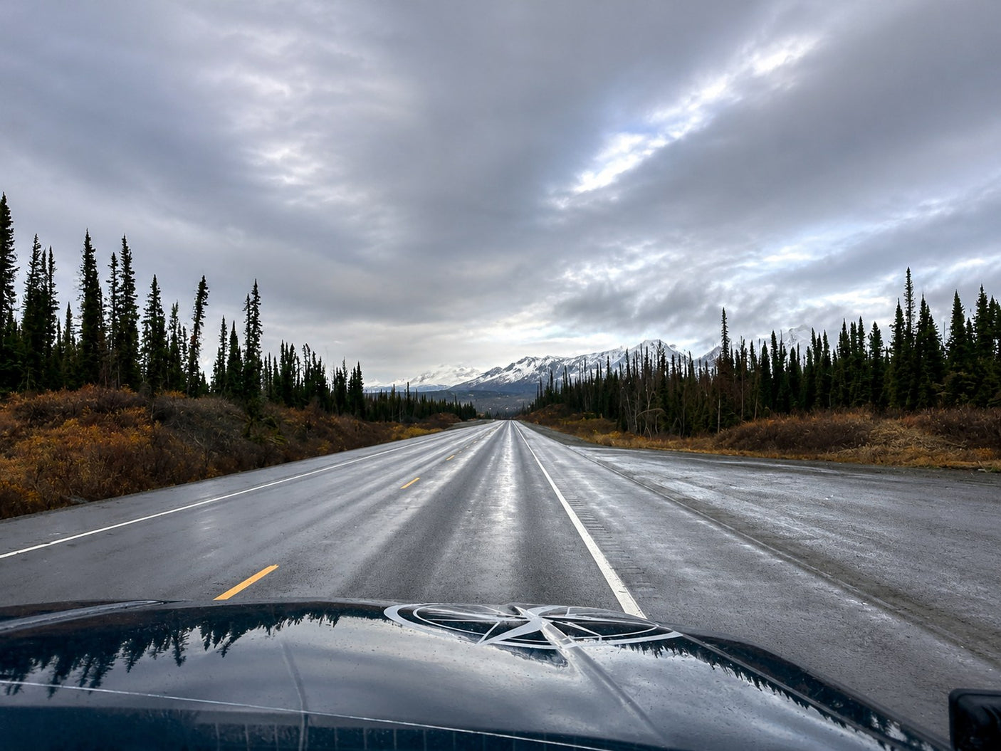 Embark on an RV rental adventure! Wet highway stretches through tall evergreens to snow-capped mountains under a dramatic, cloudy sky.
