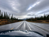 Embark on an RV rental adventure! Wet highway stretches through tall evergreens to snow-capped mountains under a dramatic, cloudy sky.