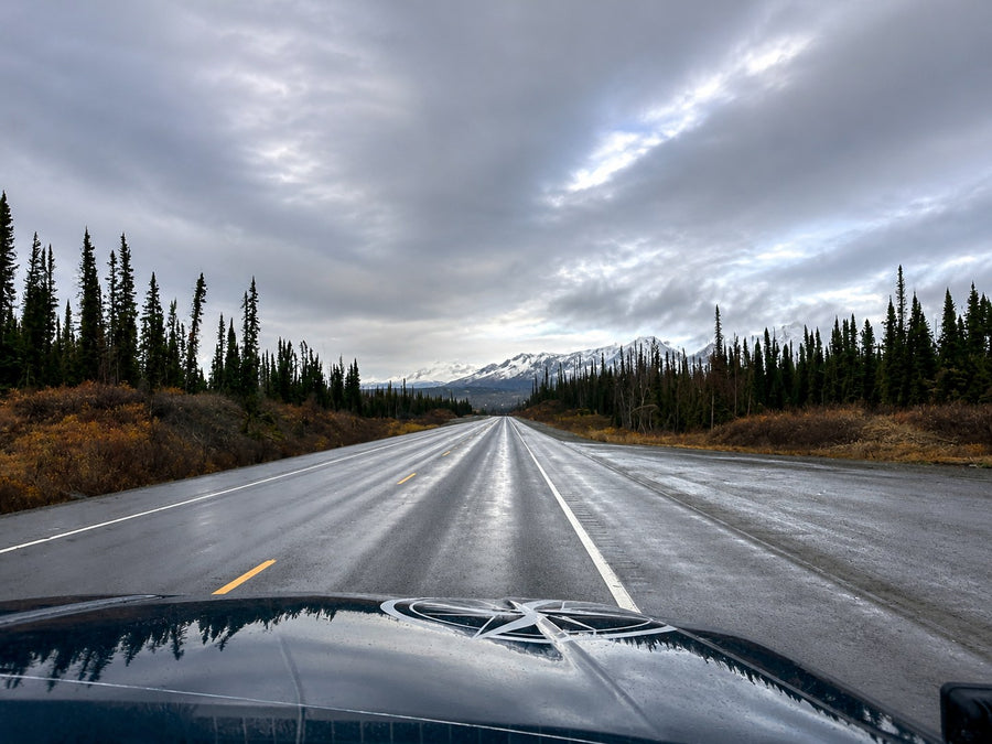 Embark on an RV rental adventure! Wet highway stretches through tall evergreens to snow-capped mountains under a dramatic, cloudy sky.