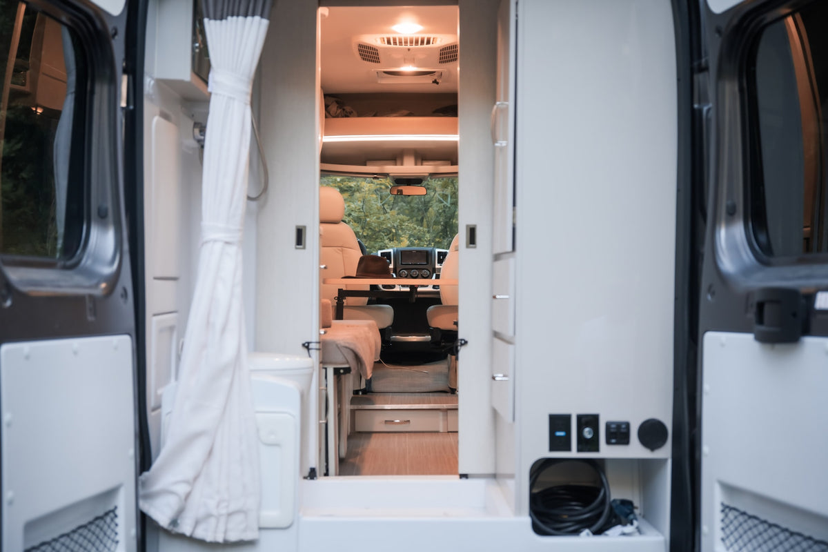 Experience comfortable rv rental adventures! View inside a camper van from open rear doors, showing the living area, bathroom, and front cab. — photo 12