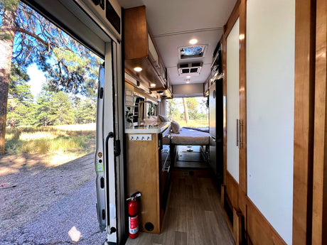 Spacious RV rental interior with kitchen, bed, and open door revealing a serene pine forest. Ready for adventure!