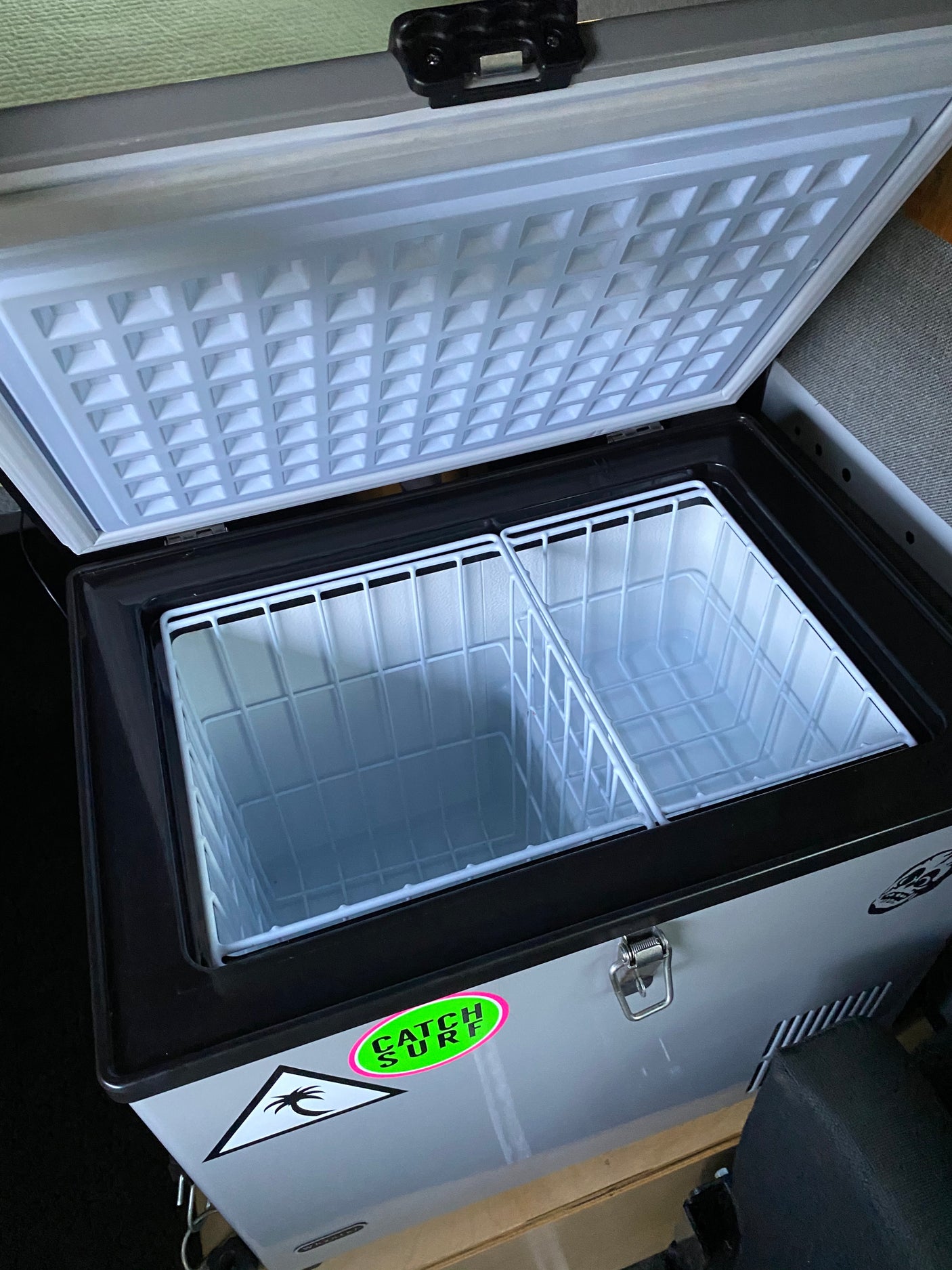 This open portable fridge/freezer, ideal for an rv rental, features two empty wire baskets & a 'Catch Surf' sticker.