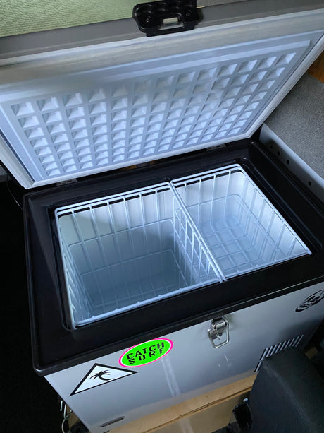 This open portable fridge/freezer, ideal for an rv rental, features two empty wire baskets & a 'Catch Surf' sticker.
