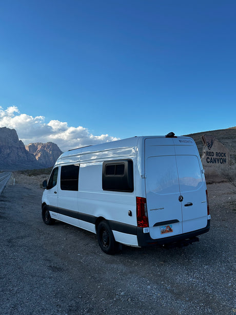 White Sprinter RV rental van parked on gravel at Red Rock Canyon, Nevada, with scenic mountains under a blue sky.