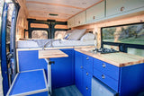 Vibrant blue interior of this RV rental camper van boasts a raised bed, full kitchen, and scenic window view.
