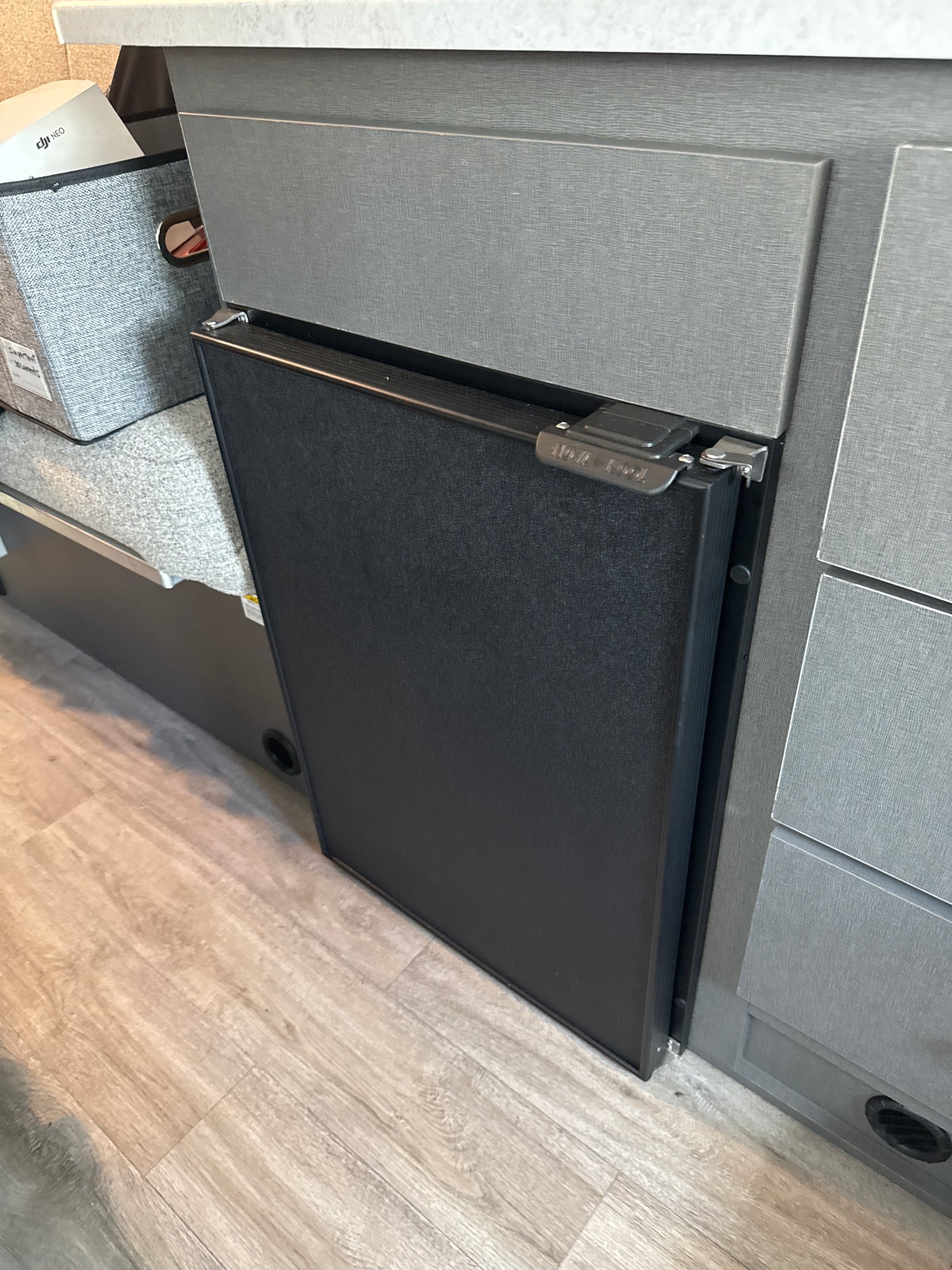 Inside an RV rental: a compact black refrigerator integrated into textured gray cabinets, next to a fabric storage bin.