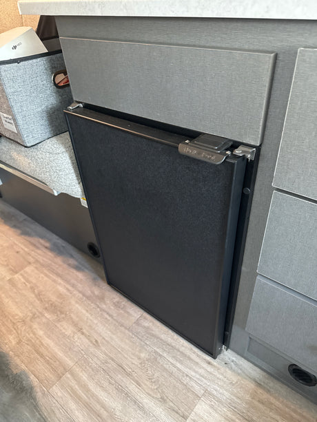 Inside an RV rental: a compact black refrigerator integrated into textured gray cabinets, next to a fabric storage bin.