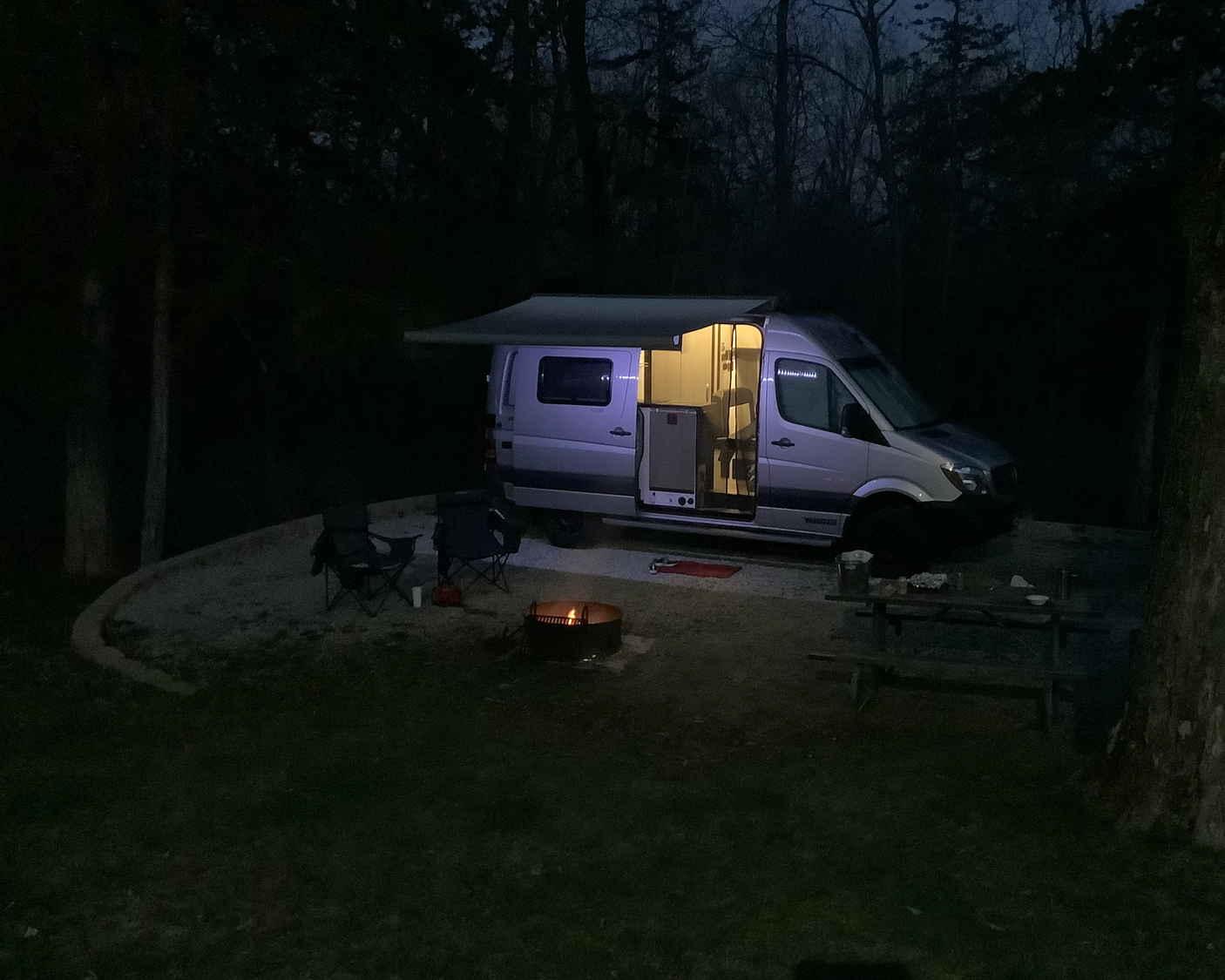 Enjoying a night at the campsite with an RV rental: illuminated camper van, cozy fire pit, chairs, and picnic table.