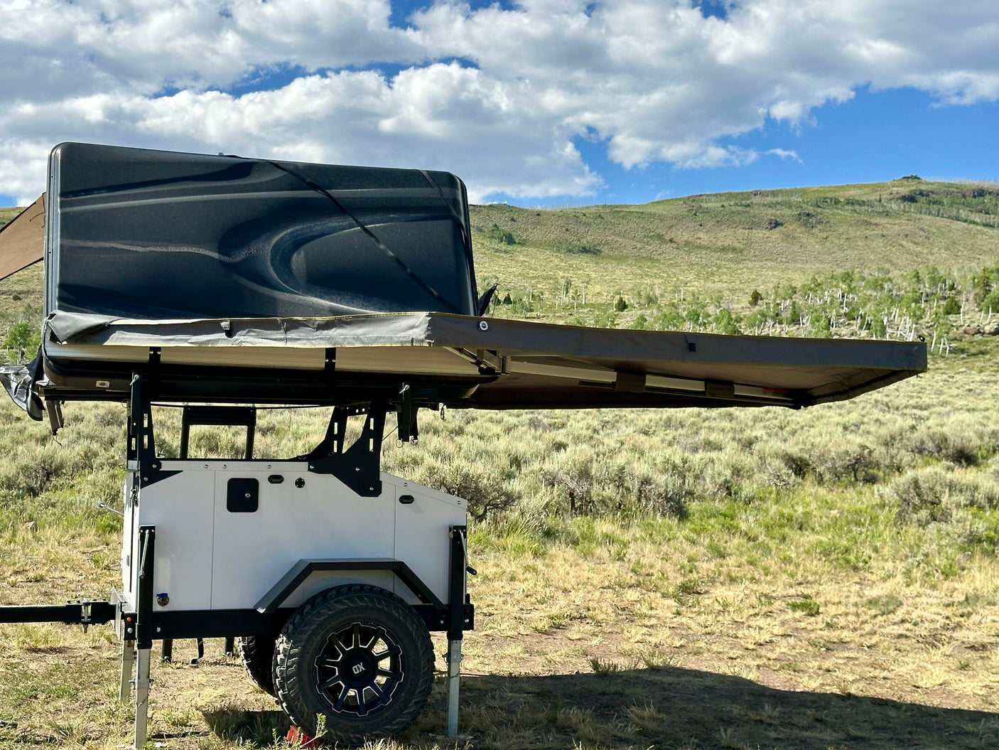 Overland trailer with deployed rooftop tent in a scenic, grassy mountain setting. A rugged RV rental option for adventure.