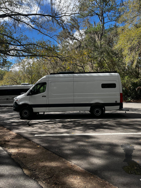 A white camper van, possibly an RV rental, parked under mossy trees on a sunny day. Blue sky, dappled sunlight on asphalt.