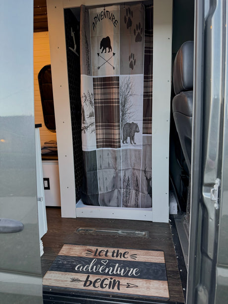 RV rental interior: A compact shower with adventure-themed curtain (bears, mountains) & 'Let the adventure begin' mat.